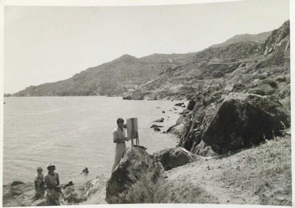 A man stands with his back to the sea on the shore of a dramatic rocky cove. He faces a canvas on an easel. Behind him is a woman wearing a hat and a young child seated on rocks by the water.