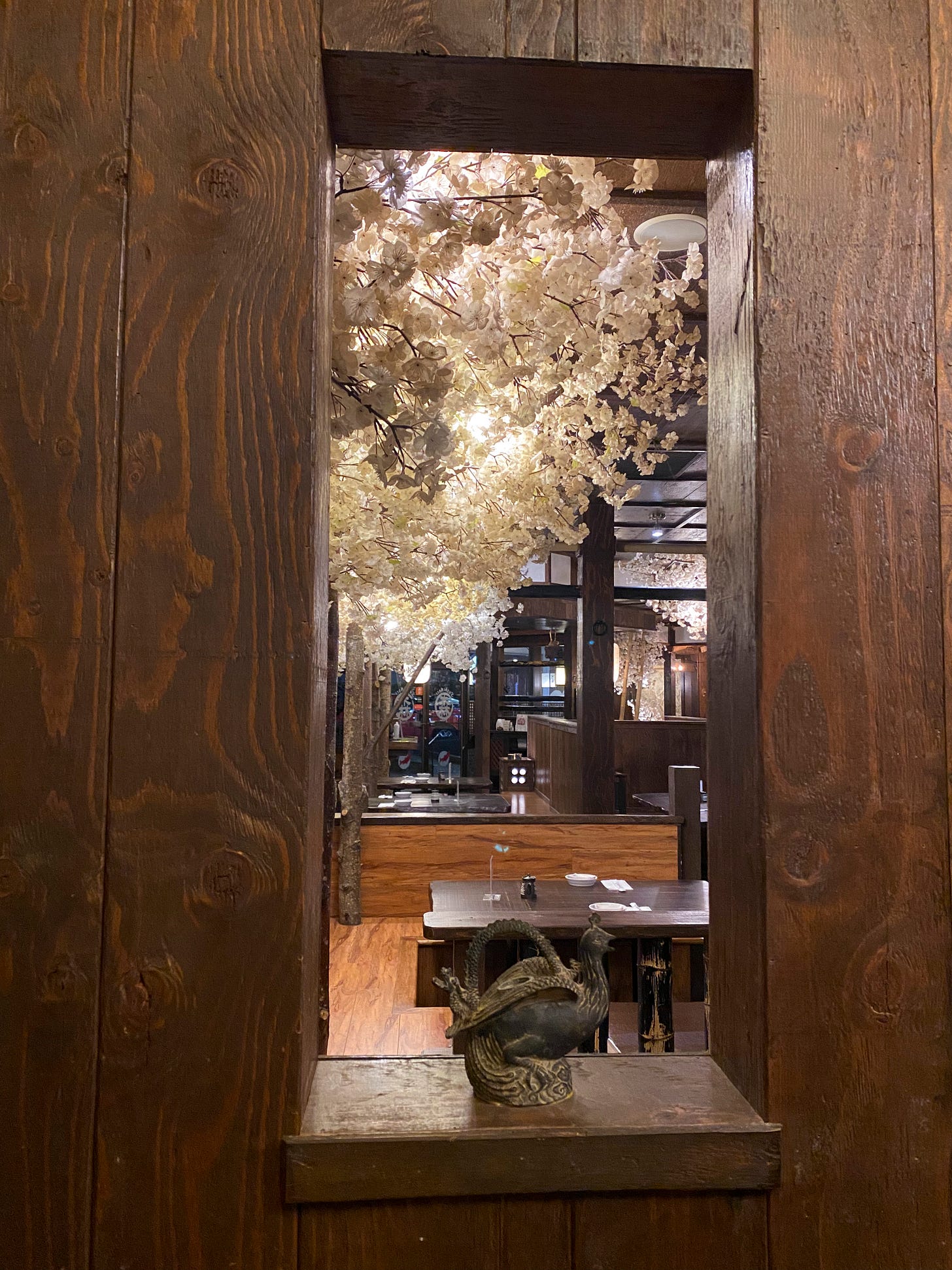 A wooden interior wall with a narrow, rectangular window showing more of the restaurant. Fake cherry blossoms surround the ceiling, lit from within. At the bottom of the windowsill is a carving of a bird.