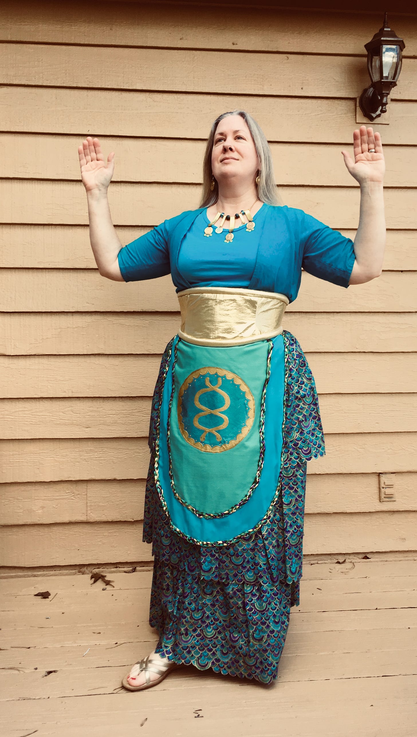 Me, a light-skinned, gray/blond-haired female-presenting person, wearing a Minoan outfit in shades of blue and gold, including a tightly fitted top, a wide belt, an apron, and a tiered skirt.