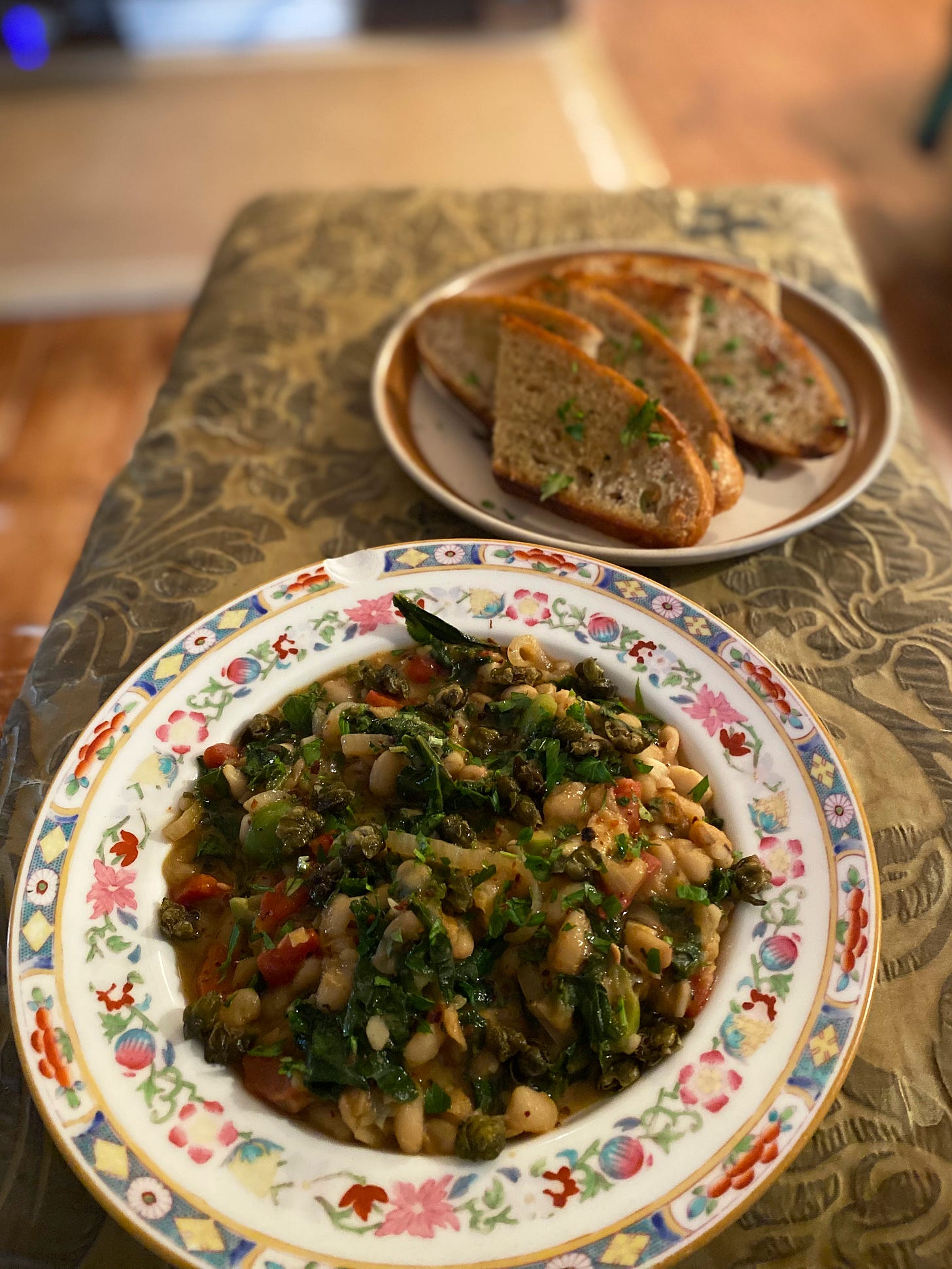 A decorated shallow bowl filled with the kale, white bean, pepper, & olive mix described above. Capers and parsley are scattered over the top. Behind it, on a mismatched small plate, slices of bread are cut in half and sprinkled with parsley.