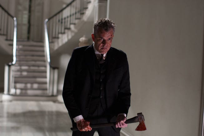 A meancing man in a dark suit holding an axe with blood splatter on his face.