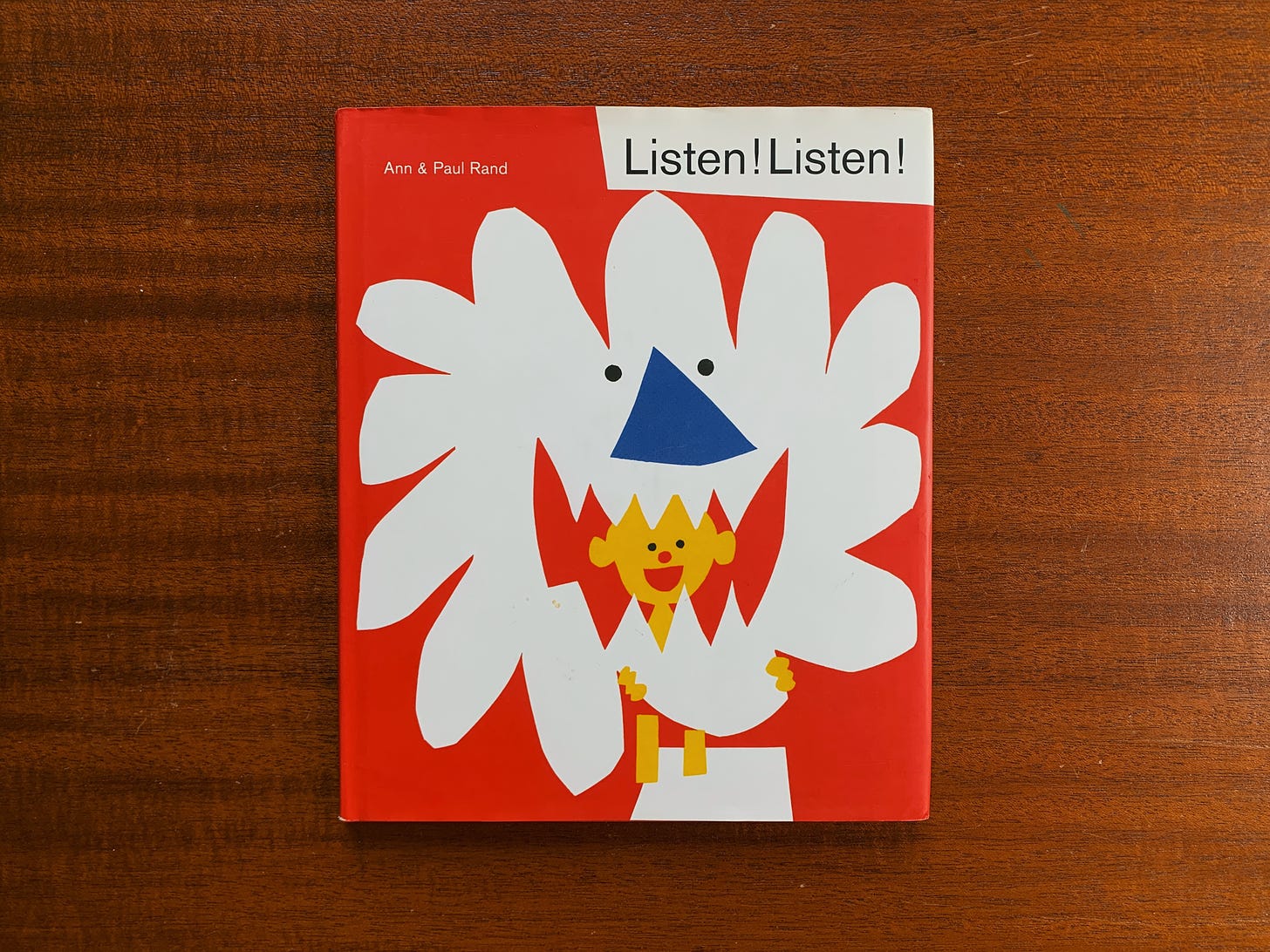 The front cover of "Listen! Listen" by Ann & Paul Rand. It's a red cover with a paper cut illustration of a child holding what appears to be a lion's face.