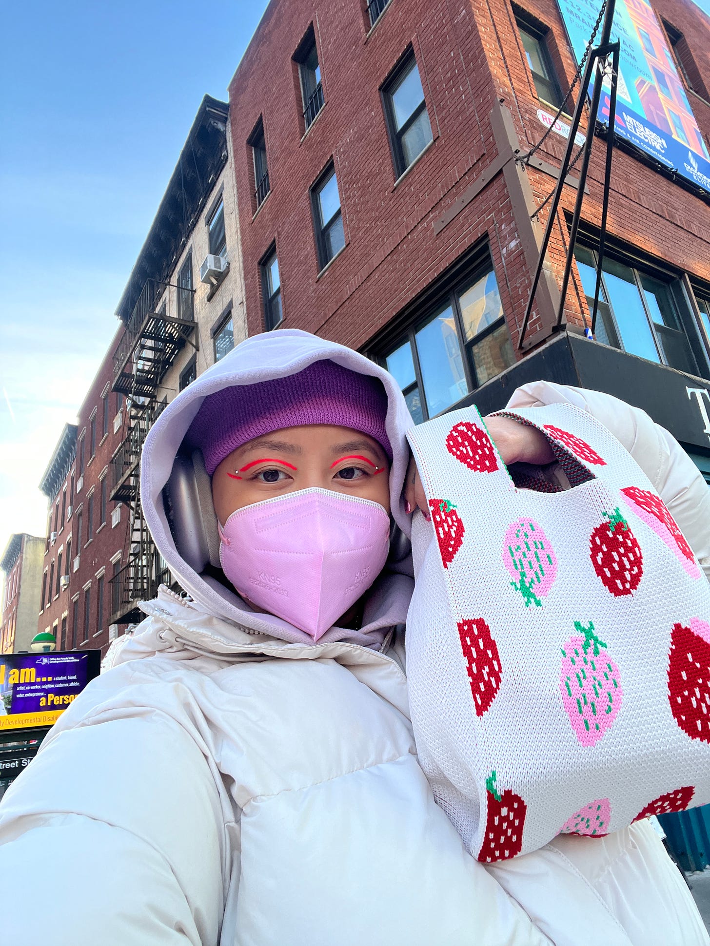 Jezz, an Asian person in their 30s, smiles with 2 KN95 masks on while outside in NYC. They're wearing a white puffer jacket, purple beanie, and holding a strawberry-print knitted tote bag. Jezz also has hot pink makeup on their eyes.