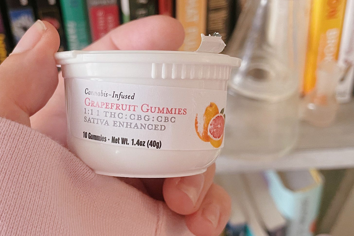 a hand holding a small container, with a label that reads "cannabis-infused grapefruit gummies, 1:1:1 THC:CBG:CBC, Sativa Enhanced, 10 Gummies, Net Weight 1.4oz (40g)" and features a picture of a grapefruit