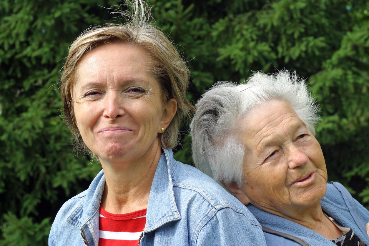 Aging white mother with gray hair rests her head on the back of her middle-aged daughter, who is also looking into the camera with a grin on her face.