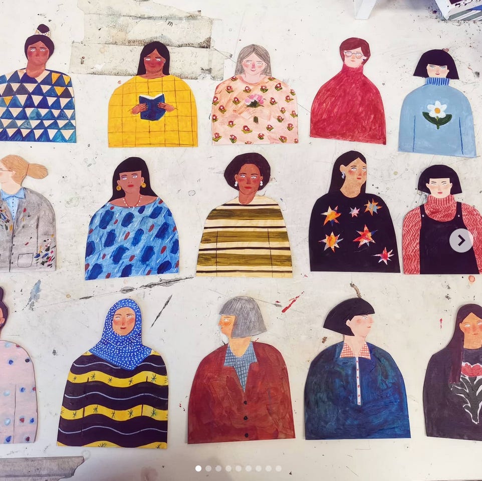 Fifteen cardboard women painted with bright colours and patterns. They siy on a very dirty desk.