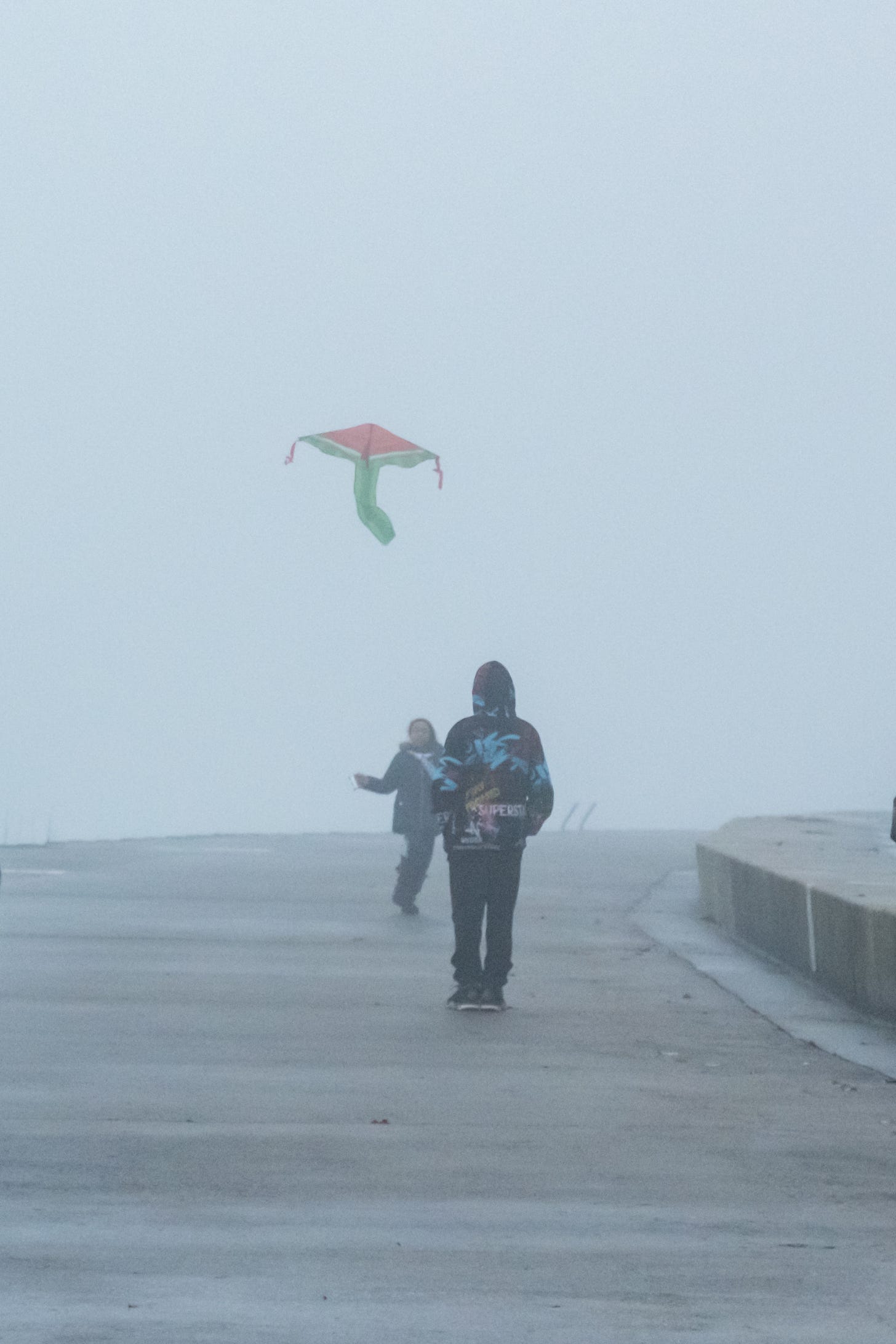 Two children try to fly a kite with the pattern of a watermelon on a foggy day with little wind.