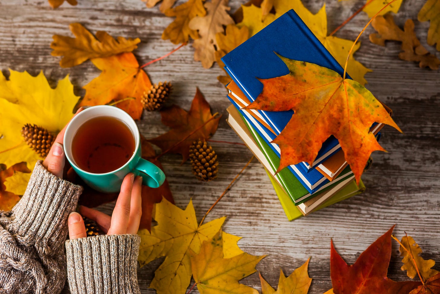 Top view of a weathered wood table, covered in fallen leaves that go from yellow to orange and red. There is a pile of colorful books to the right, and a cup of tea to the left, held by someone wearing a cozy gray sweater. 