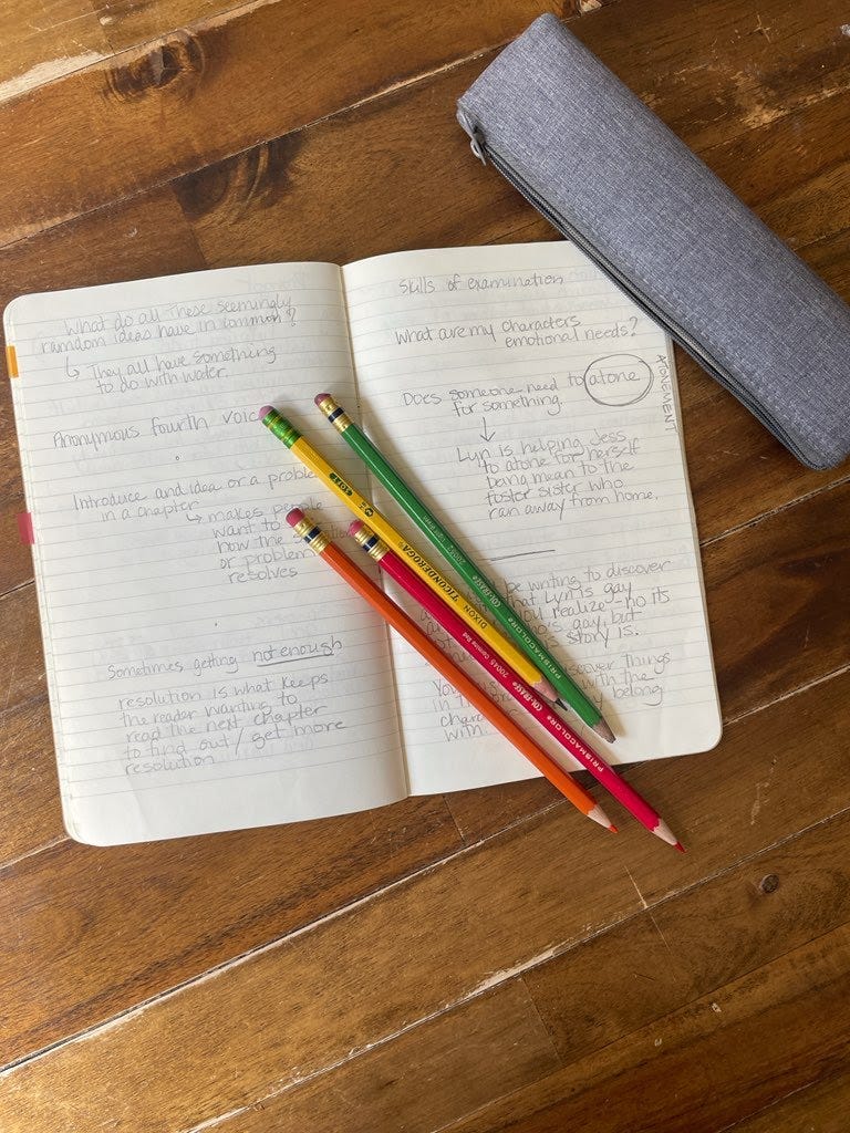 Open notebook with penciled writing inside and 4 pencils across the pages. Background is wooden table with gray pencil case