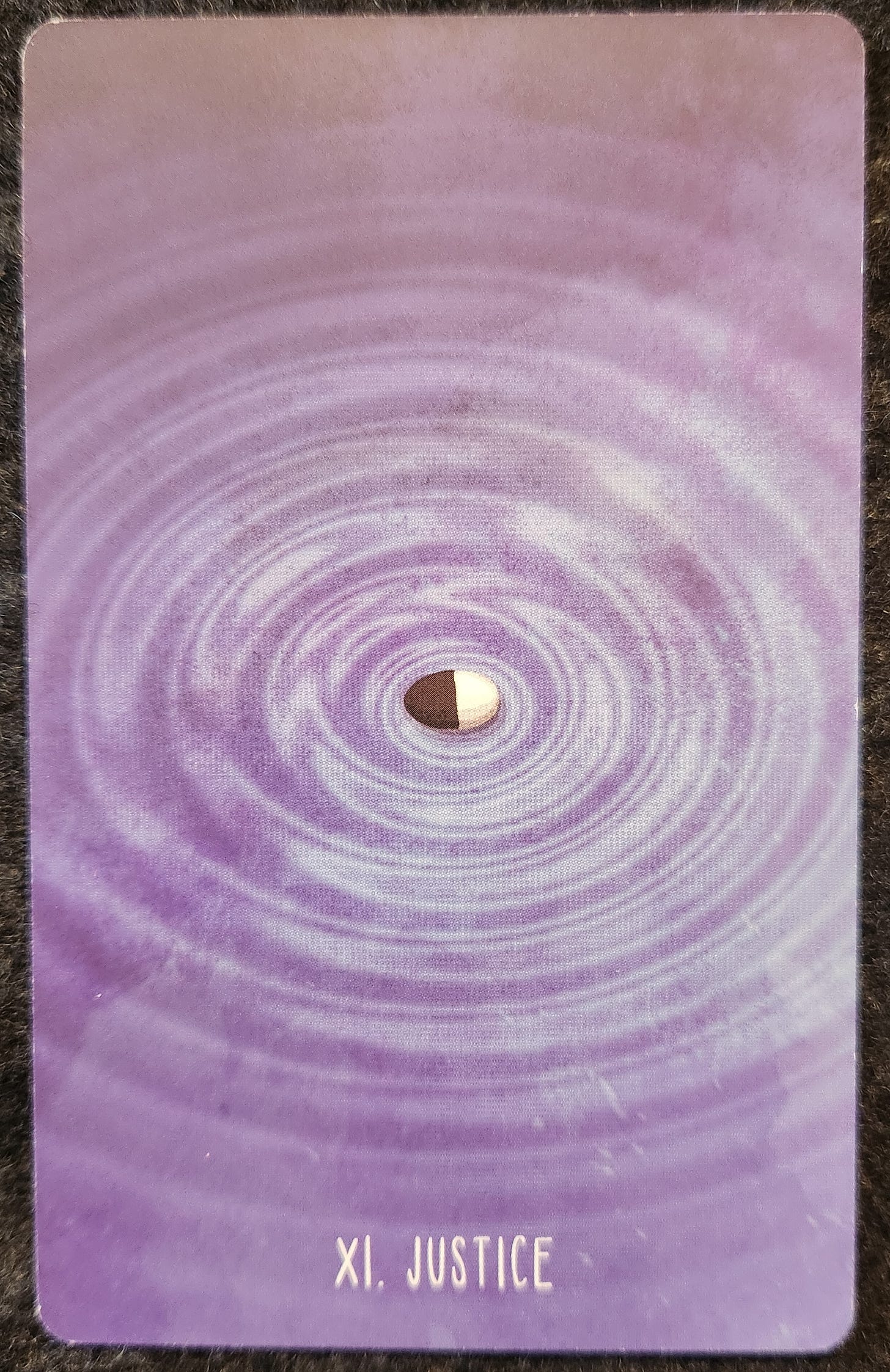 A tarot card labelled XI Justice. A pebble that is half black and half white landing on a pool of water with concentric ripples cascading out of it. 