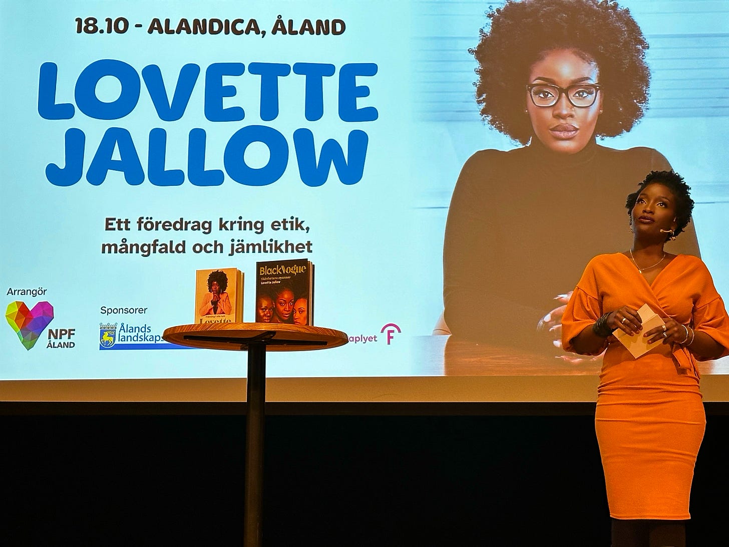 Lovette Jallow delivering a keynote lecture on ethics, diversity, and equity at Autism Åland, with her books Black Vogue and Främling i vita rum displayed on stage. Lovette Jallow delivering a keynote lecture on ethics, diversity, and equity at Autism Åland, with her books Black Vogue and Främling i vita rum displayed on stage.