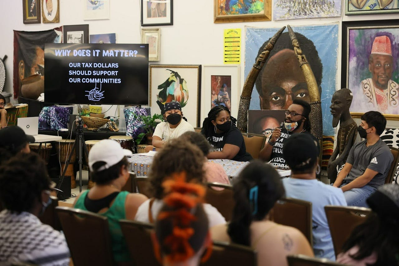 A packed room of masked organizers. Several organizers of color sit at a table, surrounded by Black artistry.