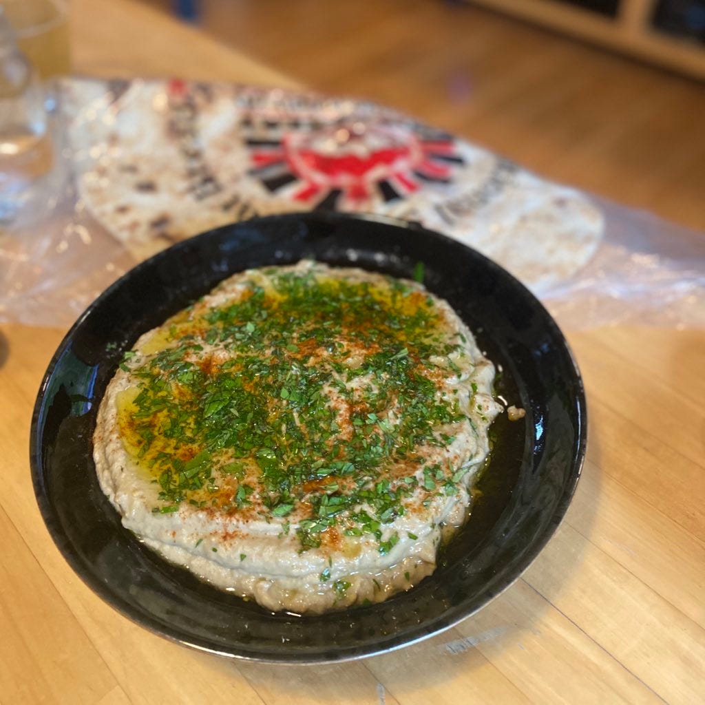 A shallow black bowl of baba ghanouj, covered with paprika, olive oil, and chopped mint & parsley. In the background is a package of taftoon.