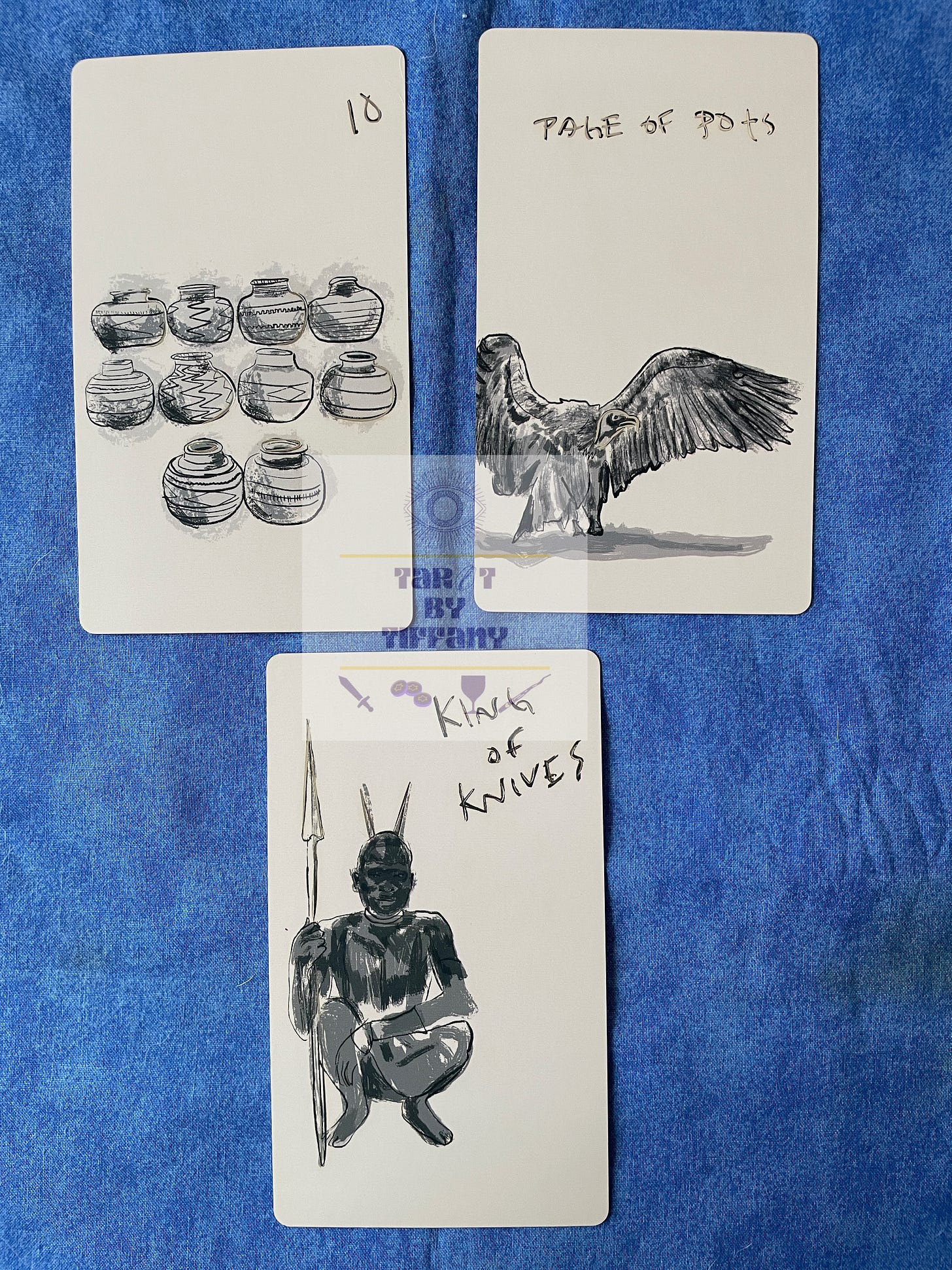 3-card reading using the Tongues of the Mother Tarot. The cards are laid out on a cobalt blue cloth background. Top two cards (L-R): 10 of Pots and Page of Pots. Bottom card: King of Knives.