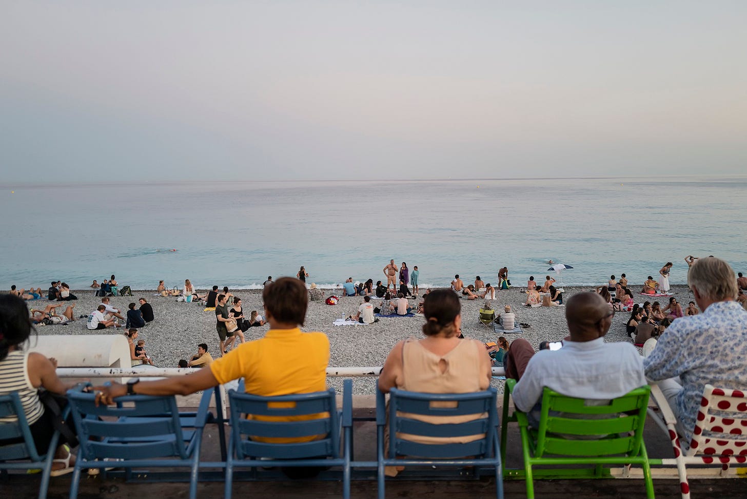 Beachfront Seating in Nice – A row of people sitting on blue chairs along the pebble beach in Nice, facing the calm, pastel-colored Mediterranean Sea. Others are scattered along the shore.