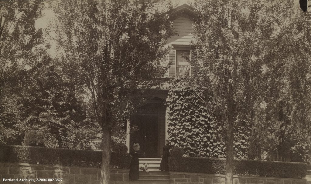 This early 20th-century photograph shows a clapboard house partially obscured by trees. Ivy grows up the walls and there are shutters on the upstairs window. Steps lead up to the porch and front door. Two women in dark clothing stand on the steps, and a medium-sized short-haired dog lies in between them.  A sandstone wall and small neat hedges are in front of the house.