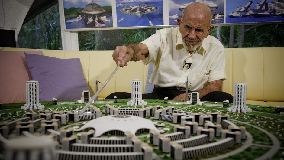 Jacque Fresco sits pointing at a model city of the Venus Project