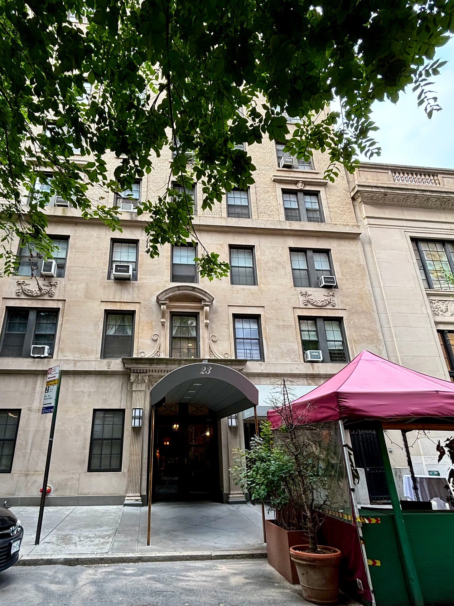 A tan brick building with a canopy of green leaves across the top of the frame. The gray awning has the address number 23. To the right is outdoor seating covered with by pink canopies for shade.