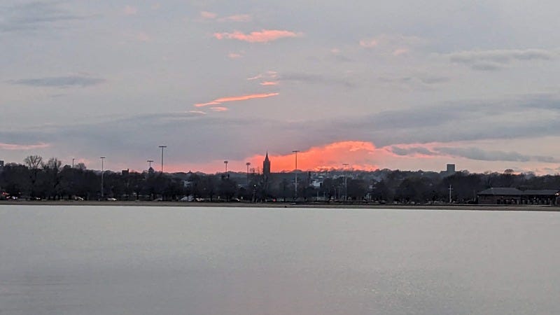 Sunset behind gray clouds with a church steeple on the horizon, over flat, calm water in the harbor