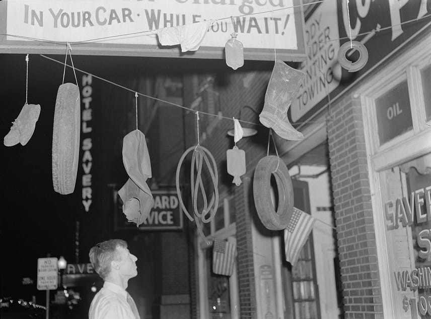 Black and white photo of the exterior of a service center. A white man looks up at rubber items hanging from ropes that have been strung up. They include a tire, rubber boots, a hot water bottle, and a hose.