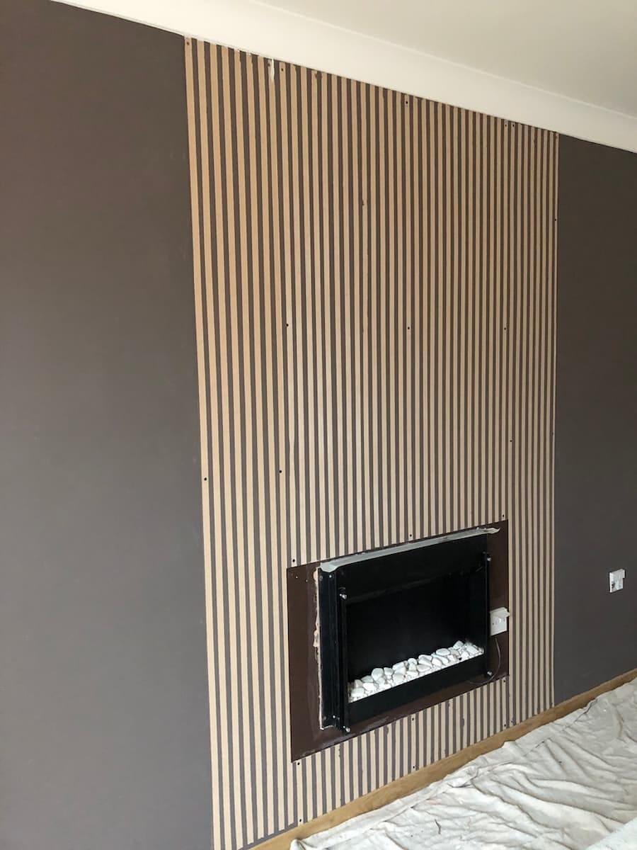 Feature wall with vertical stripes painted in the centre where wooden panels are to be positioned. The wall is painted a muted brown either side of the vertical stripes. There is a fireplace positioned centrally in the wall.