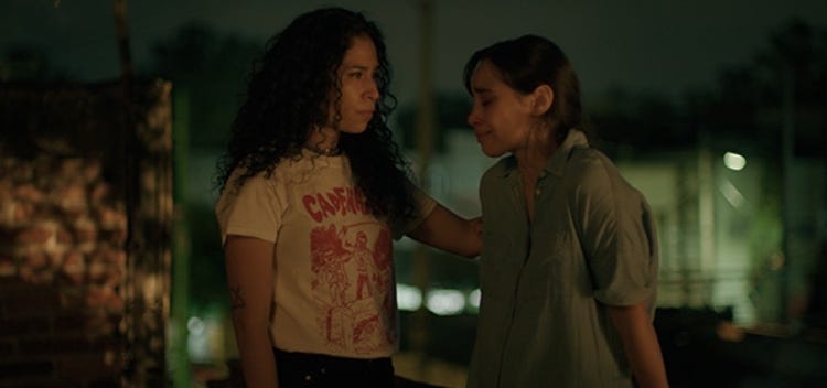 Two women stand on a rooftop in an eerily lit city. The woman on the left has long curly hair and is wearing a white band tee-shirt with red print. Her arm is lifted touching the upper back of the woman on the right, who has long hair pulled back and a light blue button-up shirt, her face is looking down and she looks upset.