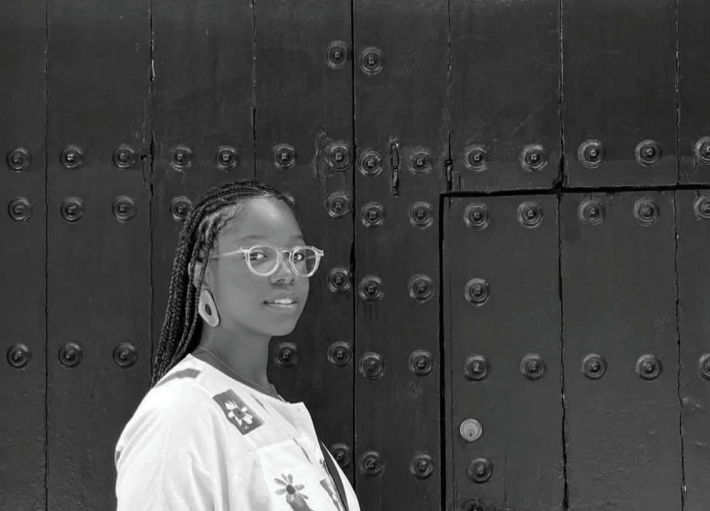 Image Description: Black and white photo of Ravon Ruffin Feliz standing in front of a black background and door wearing Fulani braids, a white shirt featuring flowers and a pair of clear eyeglass frames while staring directly into the camera with a soft gaze and even softer smile. This photo was taken by their wife on their honeymoon last year while in Spain. 
