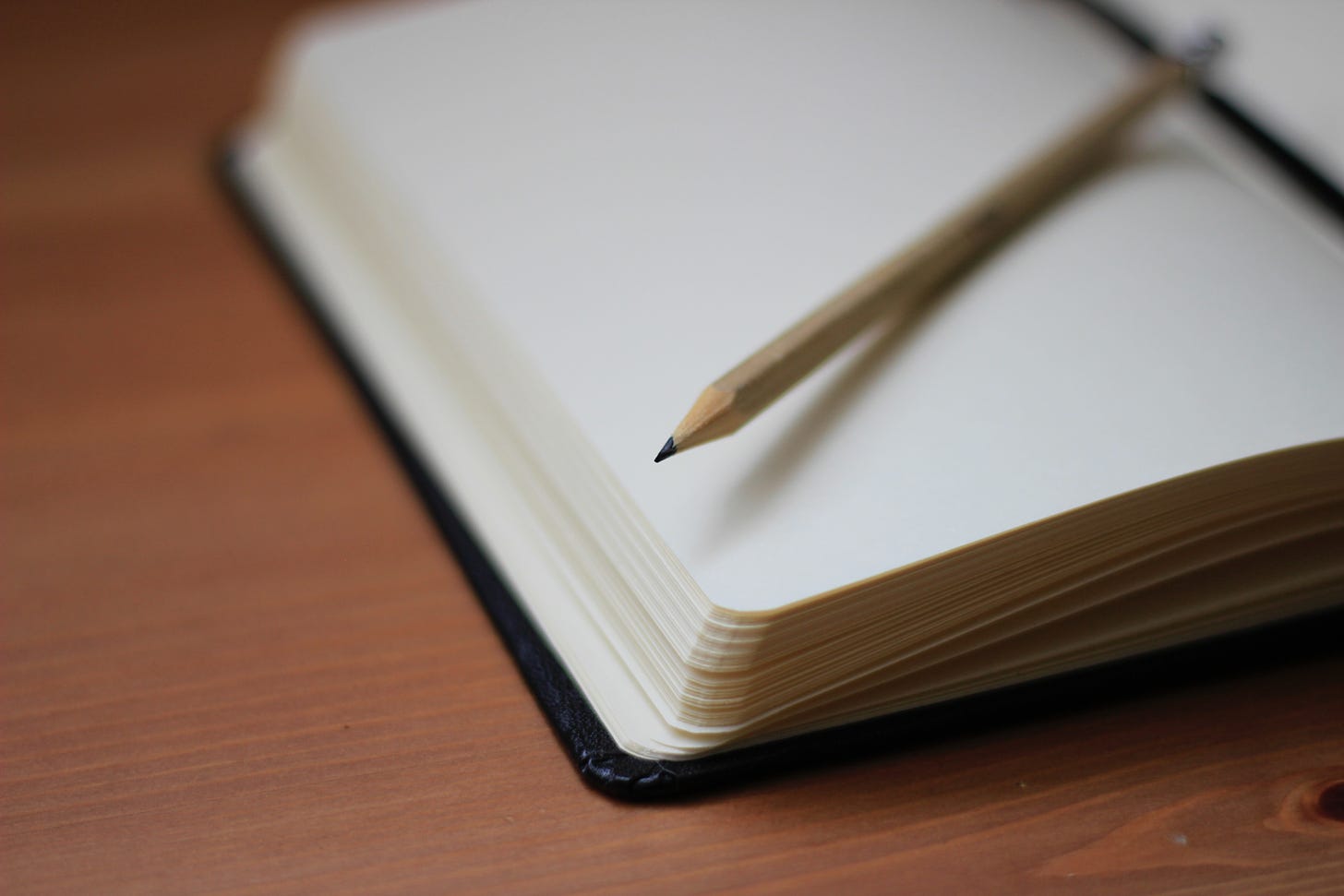 A brown pencil sits on blank white page of a notebook. The notebook is on a brown table.