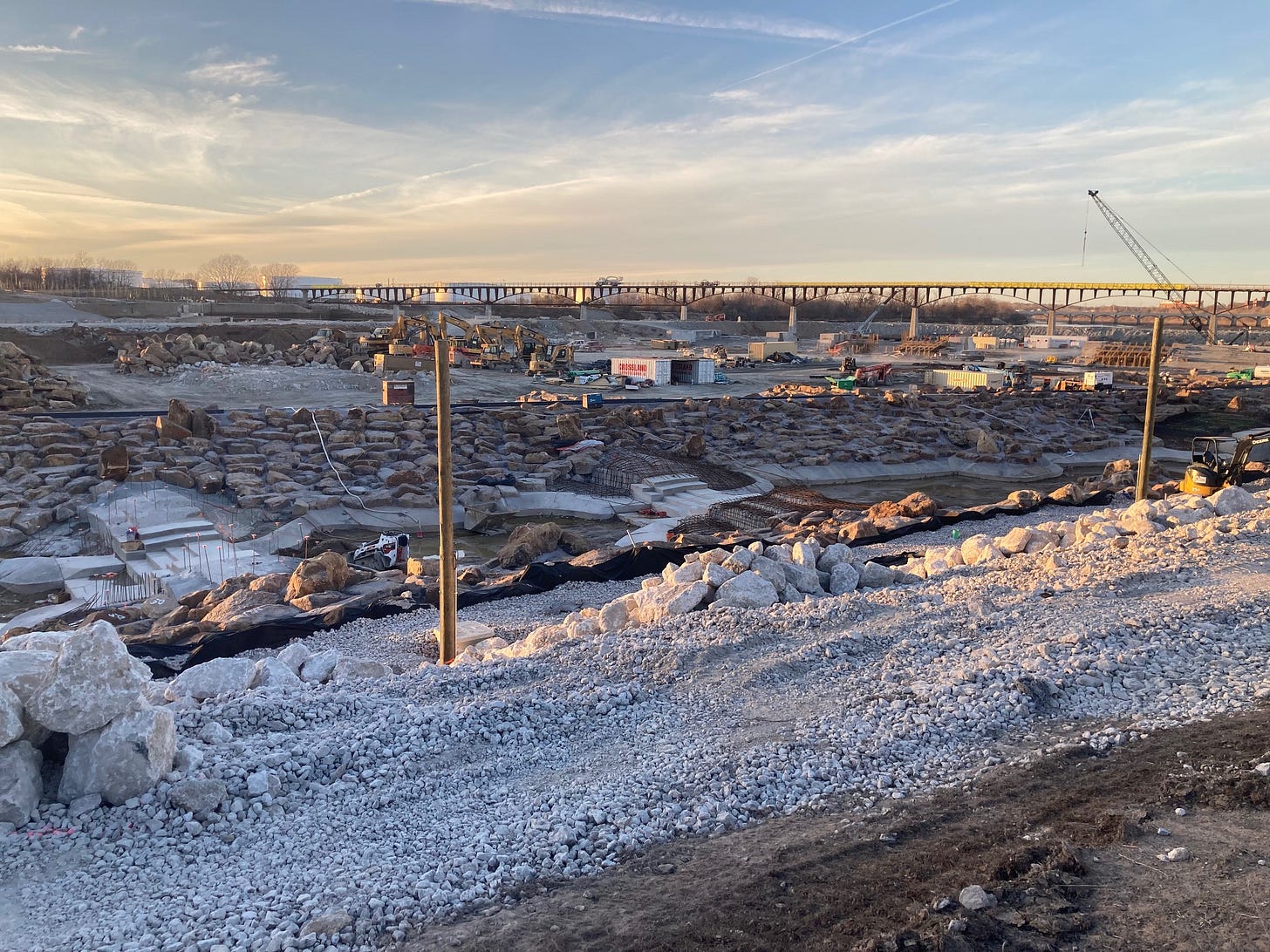 The photograph looks west as golden light glances off a vast construction pit in the Arkansas River bed beneath a cloud-streaked, blue and gold sky and refinery storage tanks on the opposite bank. The crater-like scene is filled with gravel; shipping containers; heavy machinery; and a dam, whitewater flume and bridge in progress. A sliver of river water is faintly visible a mile in the distance.