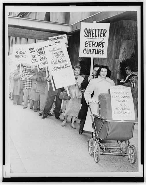 A young mother pushing a baby carriage leads a line of protesters carrying signs. One reads, "Shelter before Culture."