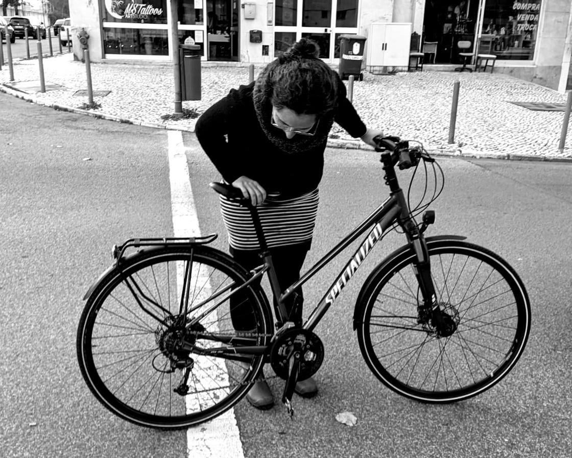 Foto em preto e branco na qual se vê uma mulher - a autora desta newsletter - e uma bicicleta preta da marca Specialized. Foto tirada em janeiro de 2023 em Lisboa.