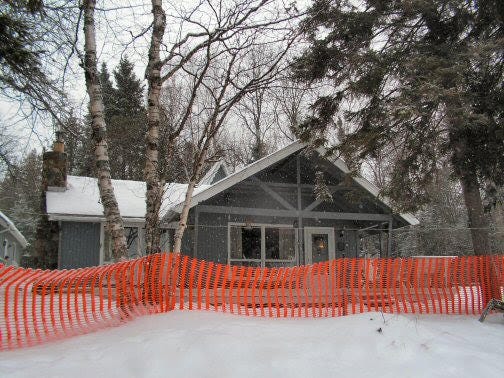 A winter cabin with snow encroaching and the bright orange snow fence doing no good at holding it back.