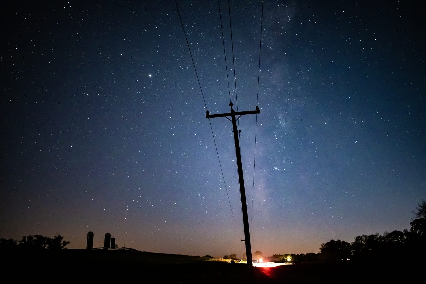 A photograph of a telephone pole and lines silhouetted against a dark, starry sky. The pole stretches vertically up through the center of the frame, following the path of the Milky Way.