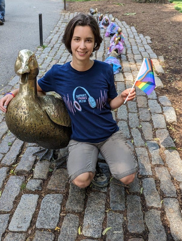 Photo of Rey and the famous duck statue in Boston inspired by the book Make Way For Ducklings. Rey is wearing a "support trans media" t-shirt and holding a progress pride flag. They have brown hair, light skin, and are smiling.