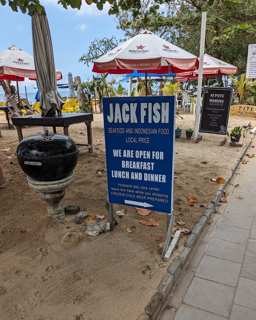 A blue sign on sand that reads, "Jack Fish Seafood and Indonesian food. local price. We are open for breakfast lunch and dinner. Husband day care center. Leave him here while you shopping"