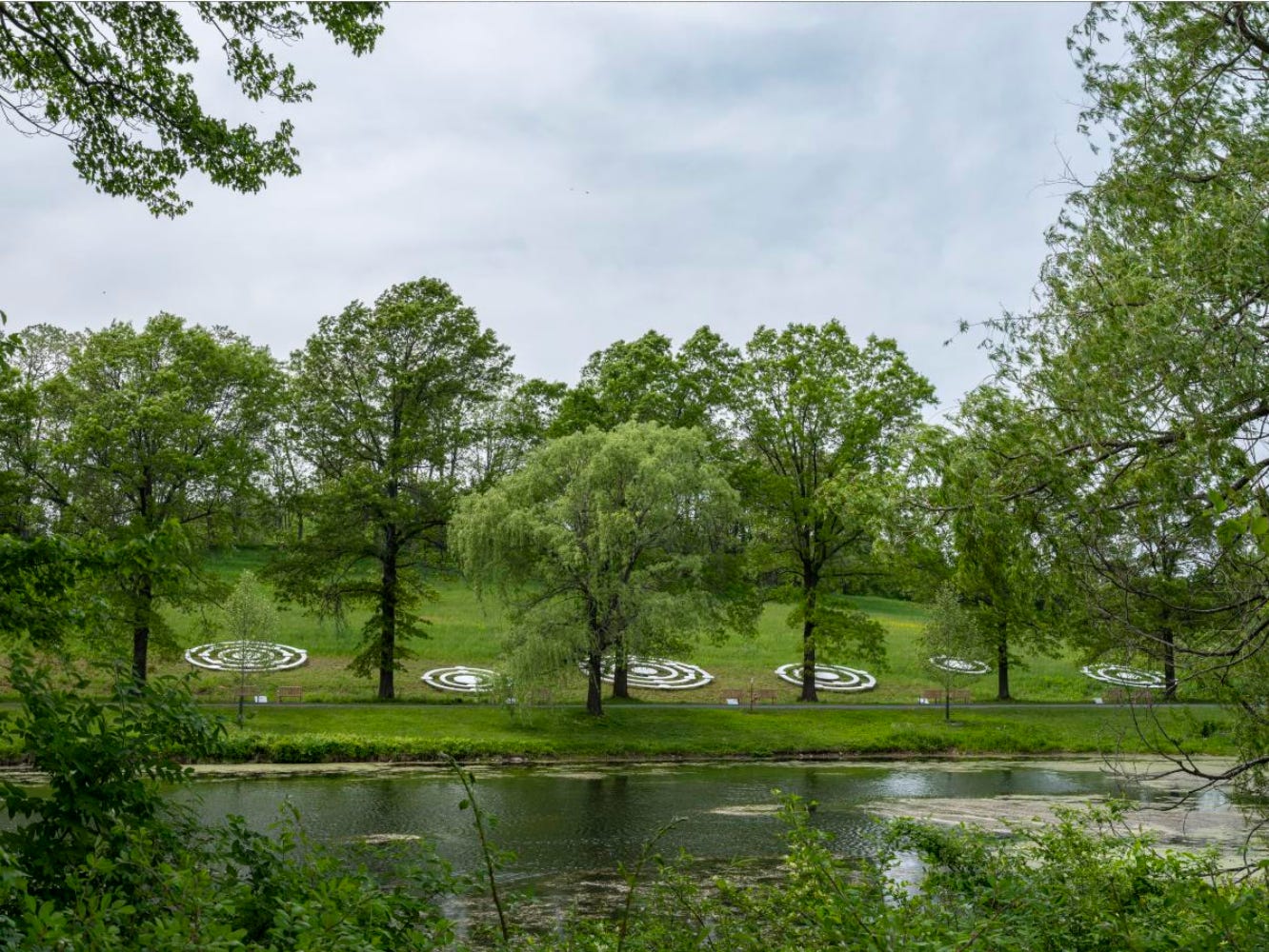 Installation view of large metal concentric circles on grass, across a lake and behind several trees.