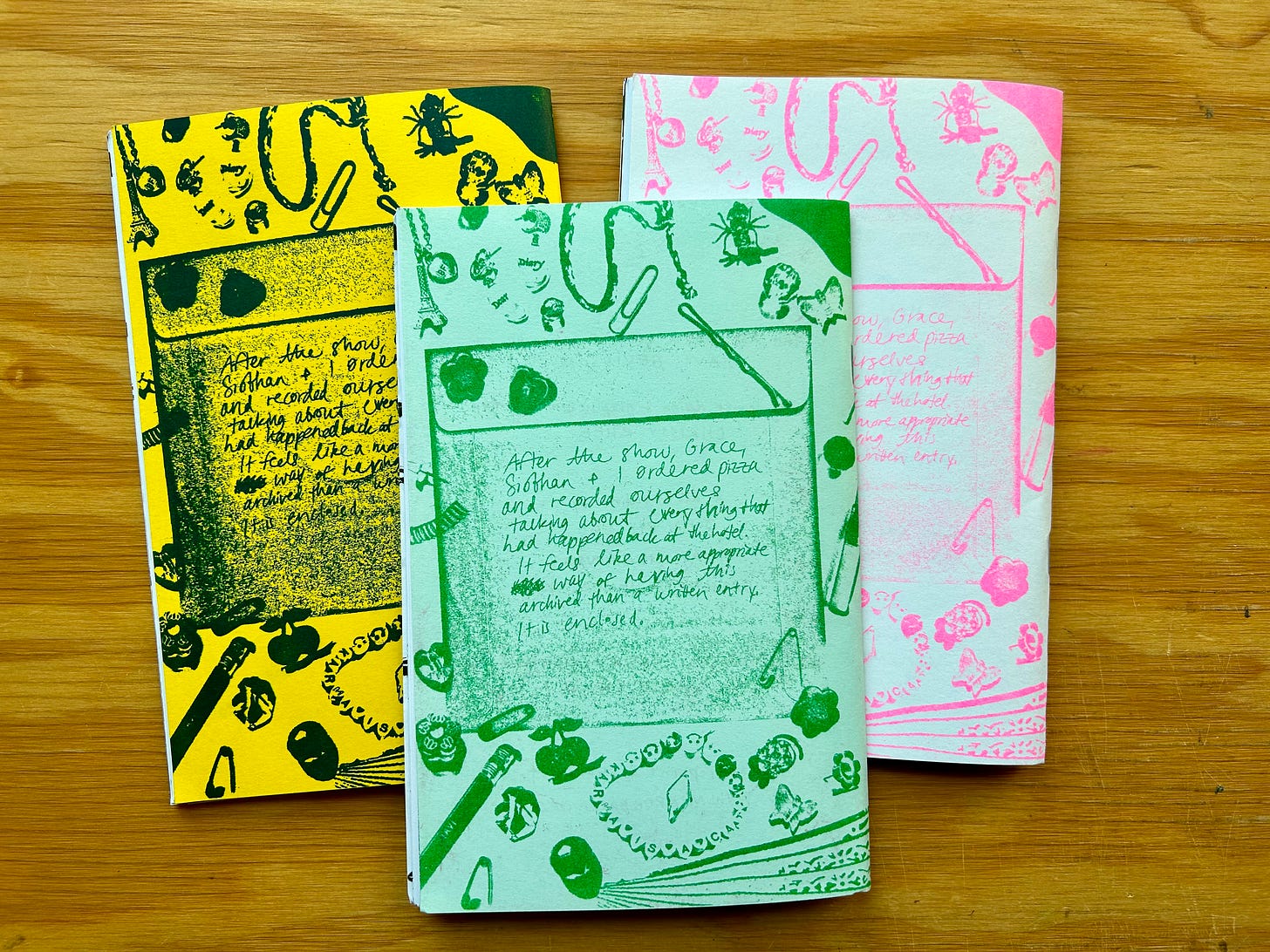 A photograph of three multi-colored copies of the zine “Fan Fiction” laid flat on a wood surface. The zines are overlapping and are turned to show the back cover. The back cover of the zine has an image of a paper CD case surrounded by a variety of small objects, which include bobby pins, bead bracelets, paper clips, flower and heart shaped beads, and two earrings. There is a hand-written note on the CD case.   The note reads:  “After the show, Grace, Siobhan + I ordered pizza and recorded ourselves talking about everything that had happened back at the hotel. It feels like a more appropriate way of having this archive than a written entry. It is enclosed.” 