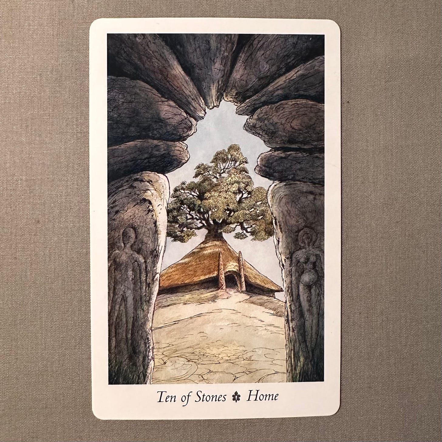 A tarot card lies on sage green fabric. The picture is bordered in white with the words Ten of Stone - Home written below it in black ink. The picture depicts the following: A great arch made up of ten weathered stones frames the picture. Through this gate we glimpse an Iron Age round house with a path leading to it. From the thatched roof of the house rises a mighty apple tree. Carved into the two lowest stones of the great arch are rough-hewn male and female figures.