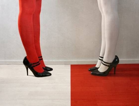 Two sets of legs knees down. One red tights, black shoes on white floor. The other white tights, black shoes on red floor.