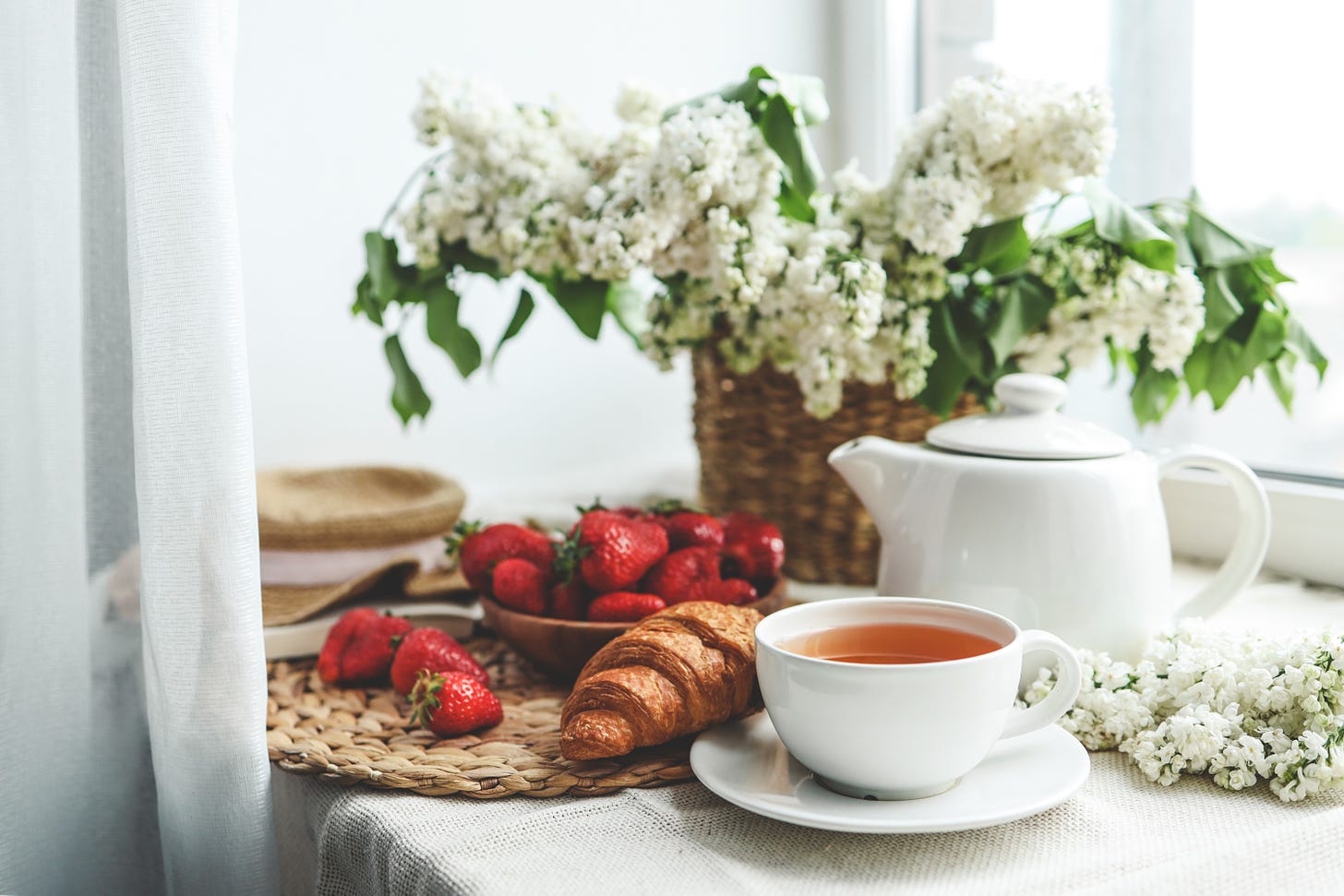 Cup of tea and teapot on a table near a window. There’s also a croissant, a bowl of strawberries and a basket filled with white flowers.