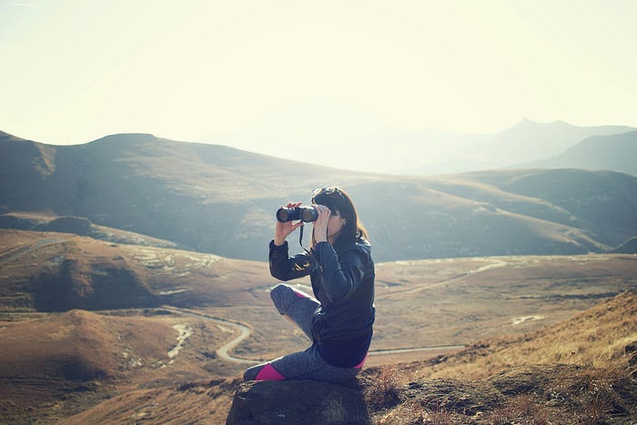 A woman sitting on a hill, looking through binoculars off into the distance with mountains behind her