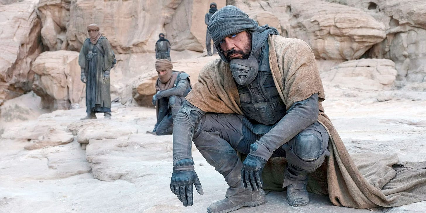 Javier Bardem as a Fremen warrior in 2021 film Dune. He's crouching on an outcrop of stone. Behind him are several other Fremen