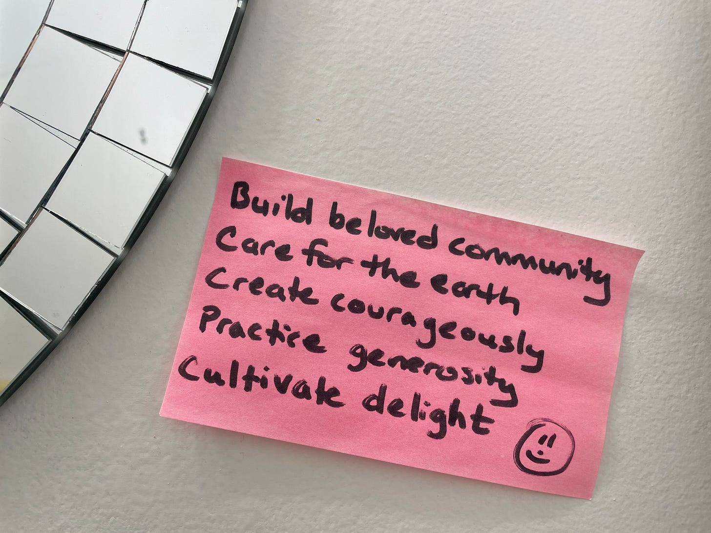 edge of curved mirror in upper right with white wall beside it. Pink sticky note. Written on it in black: Build beloved community, Care for the earth, Create courageously, Practice generosity, Cultivate delight.