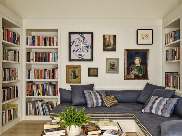 A library has a dark blue corner sofa with throw pillows and a coffee table that holds books and a plant. Books fill the shelving in the room, and seven pieces of art hang on one wall.