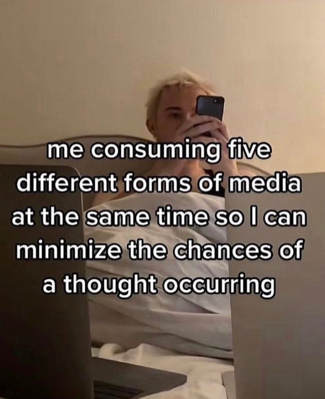 a blonde haired person sits in bed staring at their cell phone, text reads "me consuming five different forms of media at the same time so I can minimize the chances of a thought occurring"