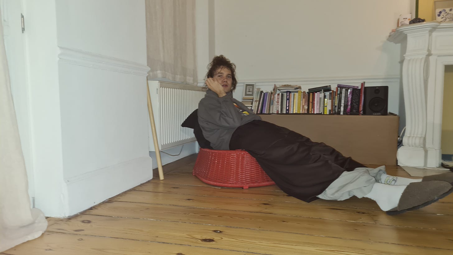 a person sitting on a red wicker chair in a room with wooden floors, white walls and a stack of books against the wall