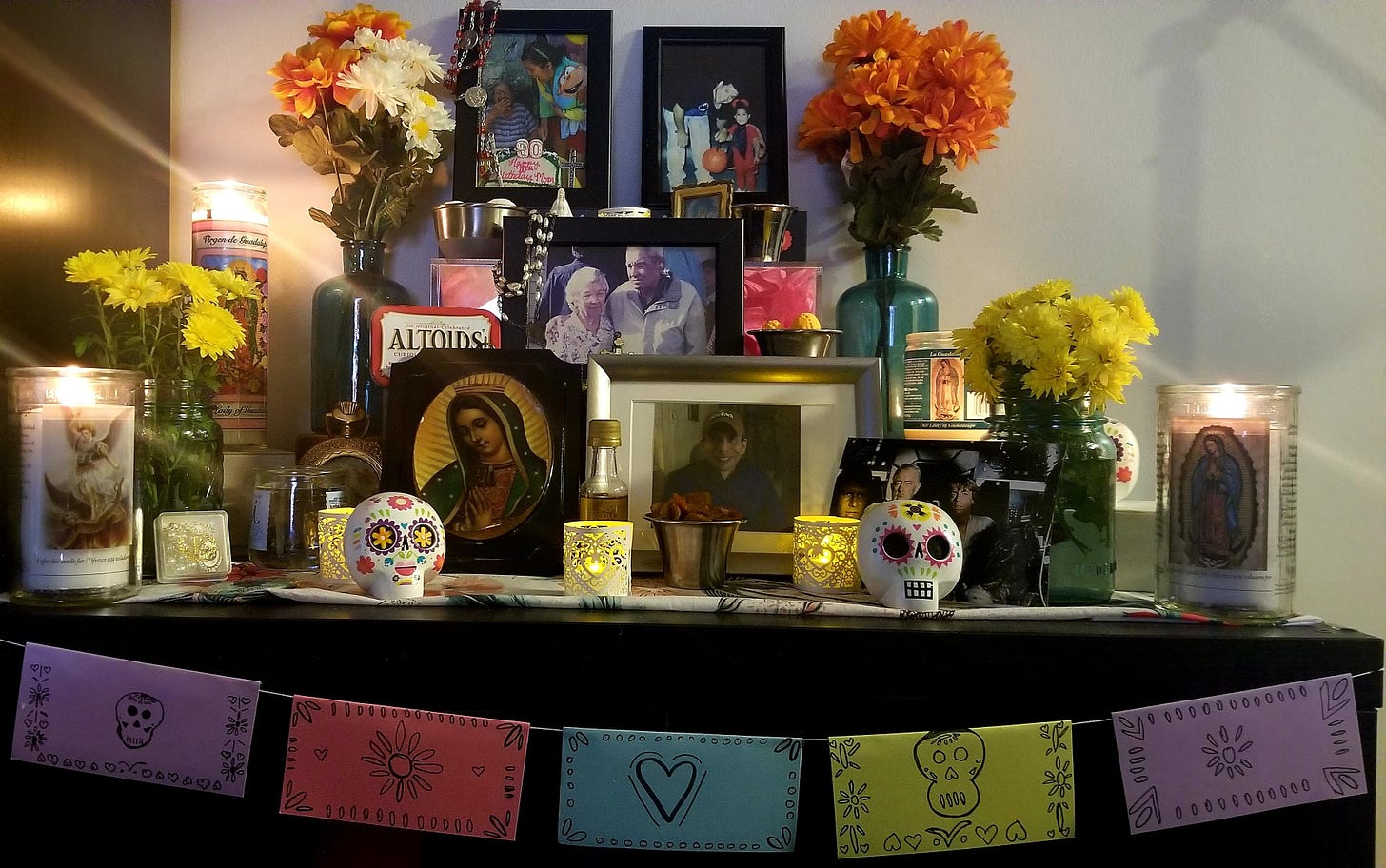 Image of ofrenda: Three tiered alter with images of Emily's maternal and paternal grandparents and Jessica's father. Calaveras, papel picada, candles with virgen de guadalupe art, and marigolds. Offerings include m&ms image of star wars movie still, altoids, whiskey, and circus peanut candy