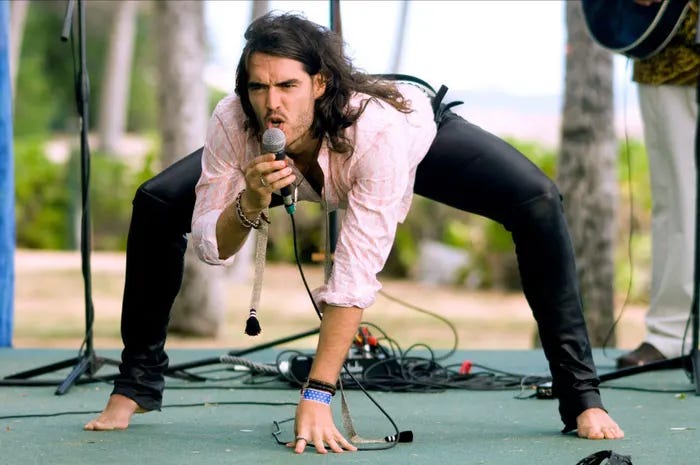 Long-haired man with brown hair and a beard wearing a pink-and-white striped shirt and black leather pants. He crouches as he sings into a microphone.