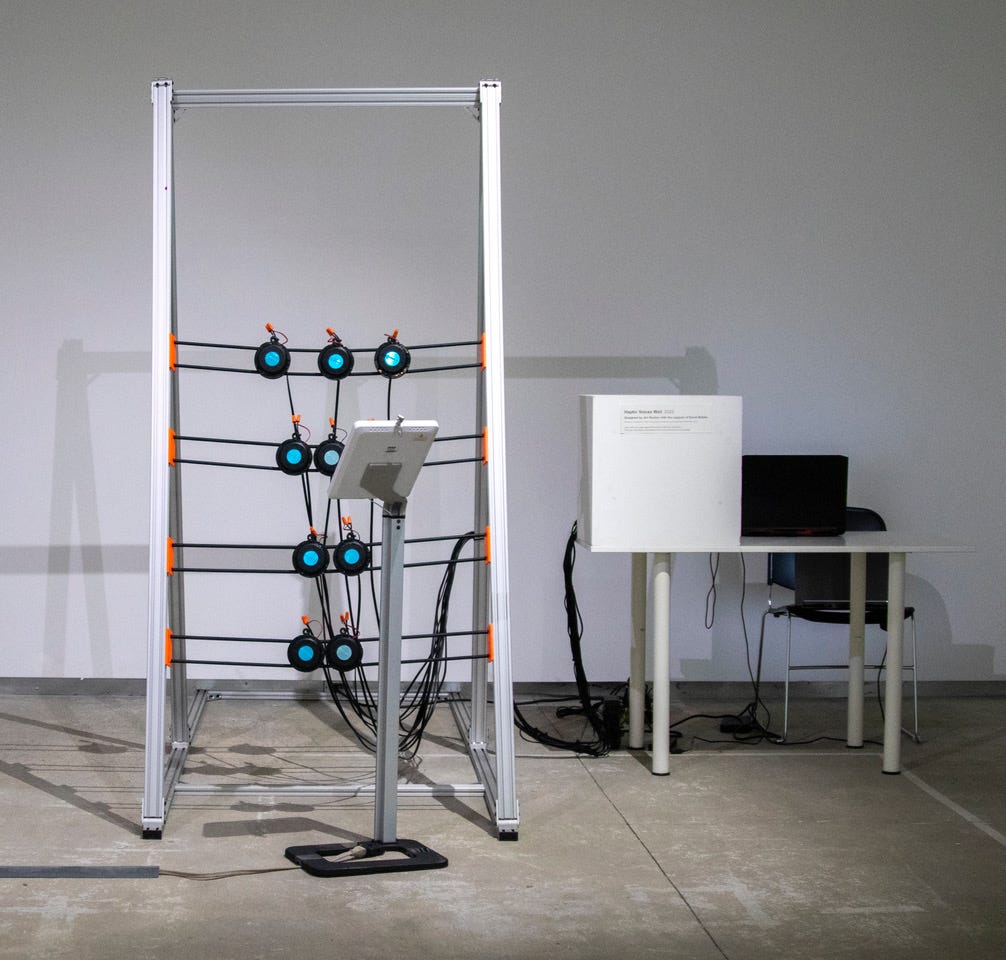 Haptic Voices installed at InterAccess. A steel frame holds 4 rows of transducers arranged to form a “wall”. To the right of the installation is a desk with a large box and computer.