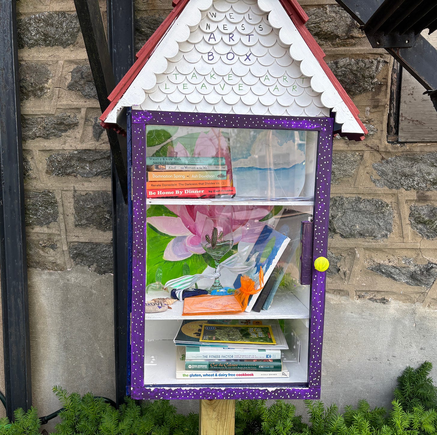 a small 'little library' house with three shelves holding bookings, paintings and small objects
